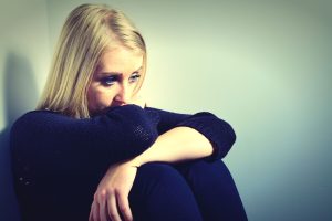 sad blond woman dressed in black sitting in a corner with arms folded. attorneys for the sexually abused help those victimized as children seek justice as adults